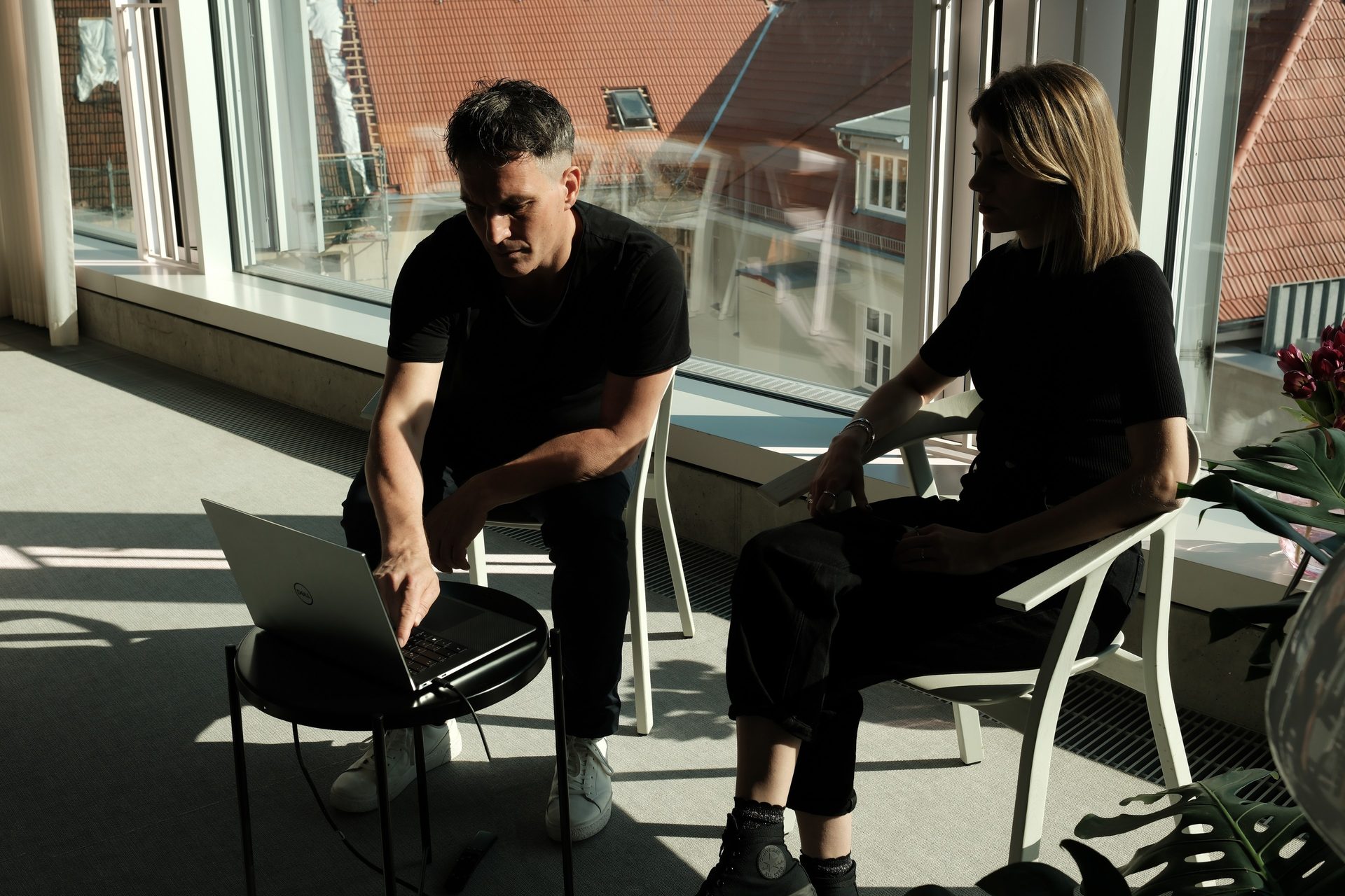 Two people in conversation at the window front, laptop on the lap, looking out over the rooftops