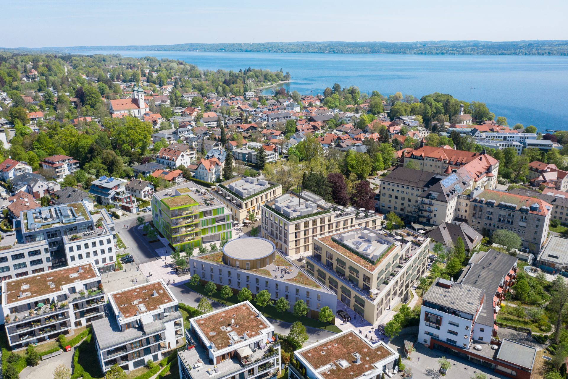 Luftbild-Visualisierung eines neuen Quartiers am Seeufer, mehrere Neubauten mit begrünten Dächern und rundem Pavillon, dahinter der alte Ortskern mit Kirchturm und der See