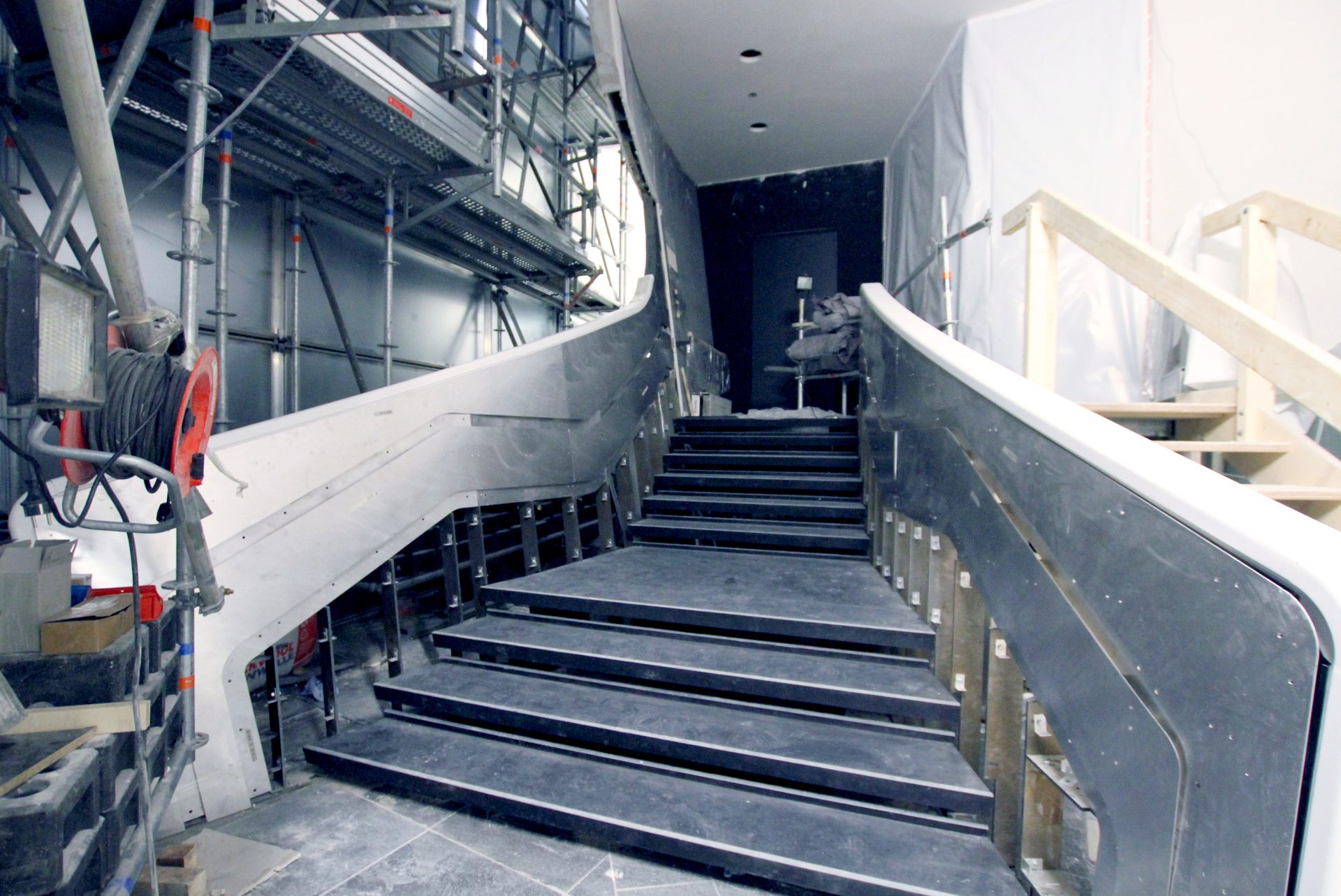 Building site with a free-form staircase, the curved balustrade and the handrail standing raw between scaffolding and dust sheeting, the treads still unclad