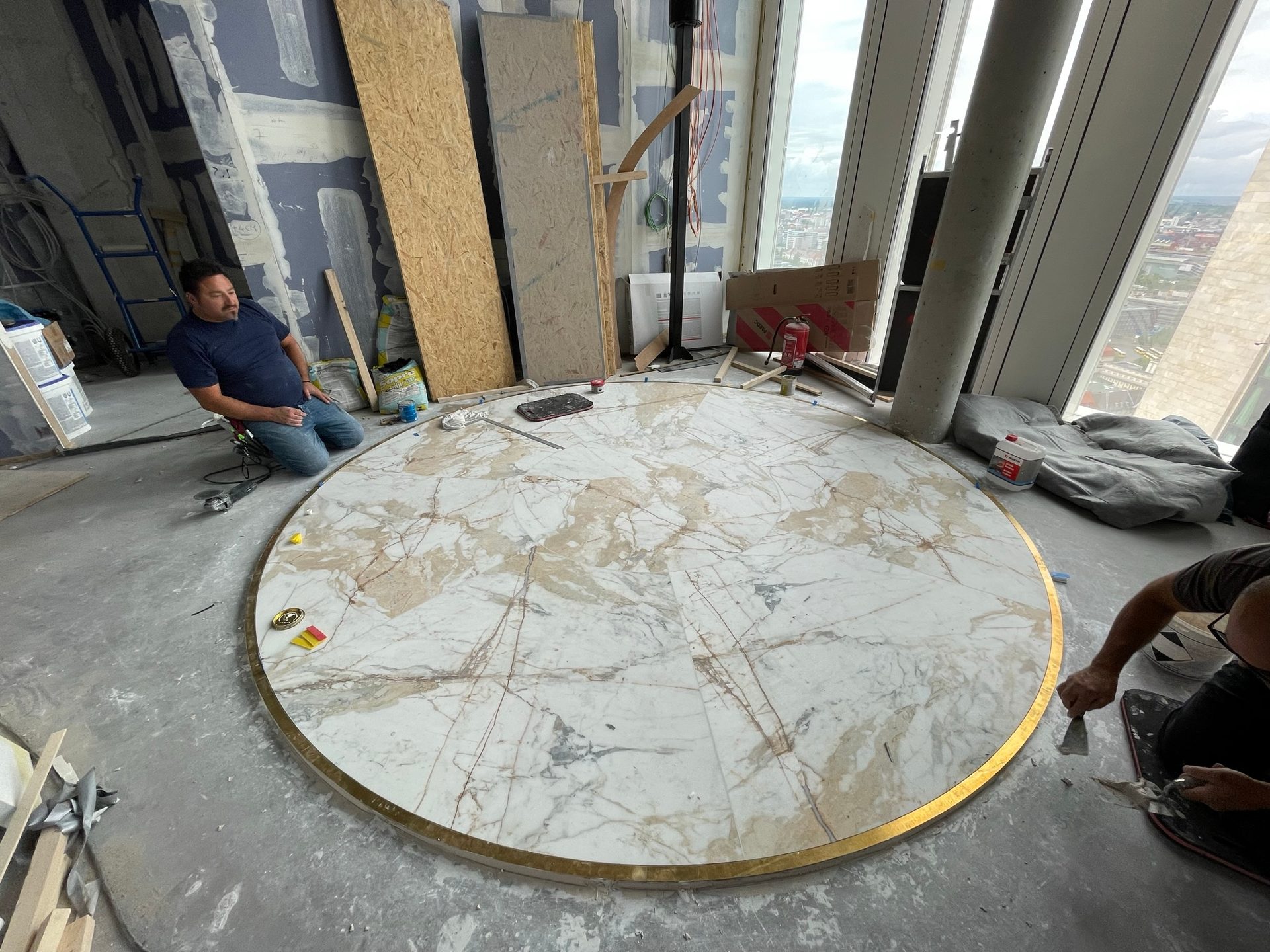 A round marble slab with a brass edge lying in the shell of a high-rise storey, two craftsmen working on it, the city behind the glazed façade