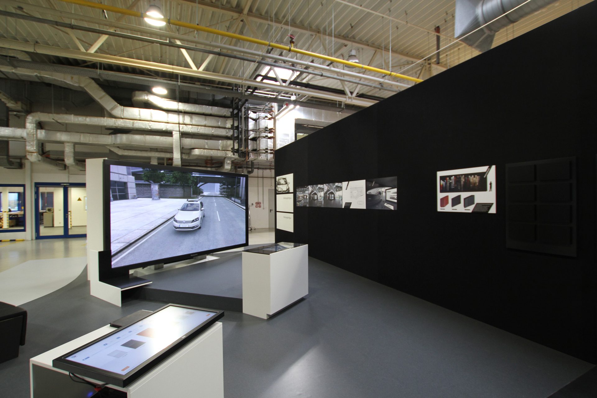 Exhibition area in a works hall, a black wall with drawings and sample boards, a large screen showing a vehicle, white display cases beneath exposed building services
